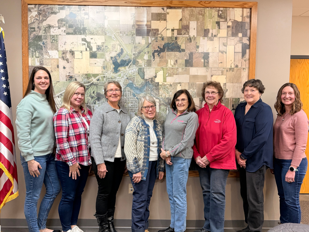 Eight women standing in front of a large aerial map with an American flag to the left.