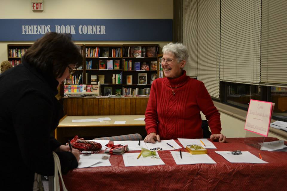 Woman in red sweater at a library's book corner, engaging with another person across a table.